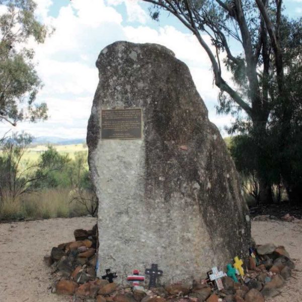 A photograph of Memorial Rock at the Myall Creek Massacre Site. A plaque is fixed onto the rock. Laid at the rock's base are small crucifixes.