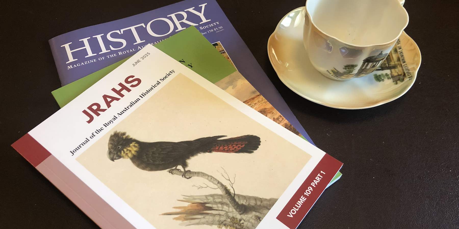 Photograph of RAHS journal and magazine beside a tea cup and saucer. The journal's cover features a picture of a black cockatoo.