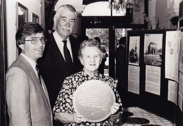 A photograph of Hazel King holding the 'History House' Green Plaque. Standing beside Hazel are Sir Roden Cutler and John Vaughan. Interpretive panels are visible in the background.