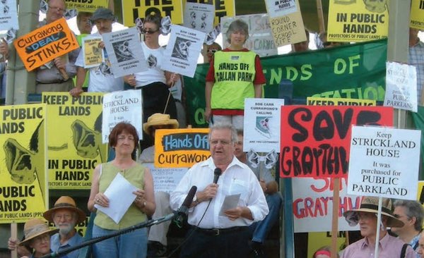 Two people stand at a microphone. Behind them is a large group of people with various protest signs, including one that reads 'Keep Strickland House - It was purchased for Public Parkland'
