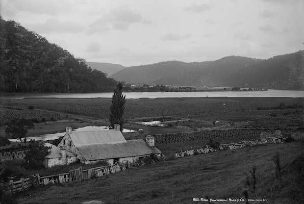 A black and white photograph featuring a farmhouse in the foreground and an undulating river in the background.