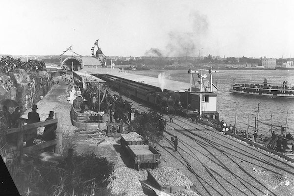 A black and white photograph of a crowd gathered to watch a steam-engine train pull into a station platform.